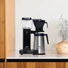 A black and silver MOCCAMASTER coffee maker sits on a white countertop, with wooden cabinets in the background. Produktbild-Vorschau