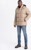 A man wearing a beige puffer jacket and blue jeans stands against a white background.