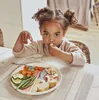 A young girl is eating at a table with a plate of food in front of her. Náhled obrázku produktu