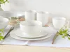 A white porcelain dinnerware set is arranged on a white tablecloth on a wooden surface, with small white flowers and green leaves as decoration. Produktbild-Vorschau