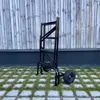 A black folding scaffold with wheels is shown outdoors, standing on a paved area with grass growing between the stones, against a wooden fence. Anteprima dell’immagine prodotto