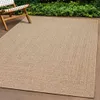 An outdoor rug with a braided pattern is placed on a patio next to a wooden chair and green bushes. Produktbild-Vorschau