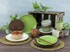 A collection of ceramic dinnerware, including plates, bowls, and mugs, arranged on a wooden table with some greenery in the background. Náhľad obrázku produktu