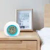 A digital alarm clock sits on a wooden nightstand next to a framed picture. Produktbild-Vorschau
