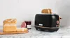 A black and copper toaster with toast in it, next to a loaf of bread and a jar of jam. Voir les images du produit