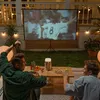 Two men are watching a movie on a projector screen in a backyard at night, with drinks and snacks on a table. Produktbild-Vorschau