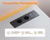 A black power strip with two AC outlets and two USB ports is shown recessed into a light-colored surface, possibly a desk or countertop. There is an orange light source in the background. Produktbild-Vorschau