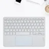 A white wireless Bluetooth keyboard with a touchpad is laid out on a white surface, with a smartphone and a cup of coffee visible in the background. Náhľad obrázku produktu