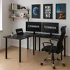 A black L-shaped gaming desk with two monitors, a laptop, a keyboard, a mouse, and an office chair in a room with shelves and artwork on the wall. Produktbild-Vorschau