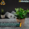 Two potted plants and bubble-wrapped items on a textured surface, with marketing text overlay. Produktbild-Vorschau