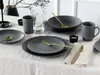 A table setting with dark gray stoneware plates, bowls, and mugs, accented with gold cutlery and a glass of water. Náhľad obrázku produktu