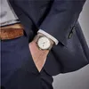A close-up shot of a man's wrist wearing a gold watch, with his hand in the pocket of a navy blue suit. Produktbild-Vorschau