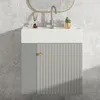 A modern bathroom vanity with a white sink and a gray cabinet with vertical ribbed detailing and a gold knob, set against a marble-tiled wall. Produktbild-Vorschau
