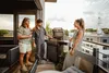 Three people are gathered on a balcony, with two men standing near a gas grill and a woman standing to the side, all appearing to be enjoying a barbecue. Produktbild-Vorschau