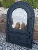 A black cast iron oven door with a glass window is placed outdoors on gravel and rocks. Náhľad obrázku produktu