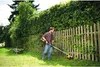 A man is using a string trimmer in a garden next to a wooden fence and a hedge. Produktbild-Vorschau