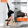 A woman is performing a yoga headstand on a wooden yoga stool with a thick black cushion, leaning against a white wall. There are plants in the background and a yoga mat on the floor. Voir les images du produit