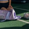 A person is kneeling next to a purple duffel bag and a pair of sneakers on a green turf surface. Produktbild-Vorschau