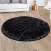 A round black shaggy rug is placed on a wooden floor in a living room setting, with a white sofa and a rattan chair visible in the background. Anteprima dell’immagine prodotto