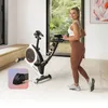 A woman is exercising on a stationary bike in a bright, modern room with a large window overlooking a garden. Produktbild-Vorschau