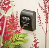 A black key safe with a 4-digit combination lock is mounted on a textured cream-colored wall, partially obscured by vibrant pink astilbe flowers in the foreground and right side. Produktbild-Vorschau