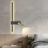 A modern wall lamp with a black and gold finish is mounted on a textured gray wall above a wooden headboard and nightstand. Anteprima dell’immagine prodotto