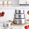 A set of stainless steel food containers with clear lids are stacked on a wooden cutting board, with some tomatoes and a lemon slice nearby. Produktbild-Vorschau