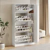A white shoe cabinet with multiple shelves and doors, filled with shoes and some accessories, placed in a room with a plant and a sofa in the background. Náhľad obrázku produktu