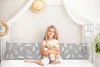 A young girl sits on a bed holding a teddy bear, with a large pillow behind her. Produktbild-Vorschau