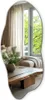 A modern living room with a large, irregularly shaped mirror reflecting a window with lush green trees outside. Podgląd zdjęcia produktu