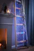 A wooden ladder decorated with string lights and greenery, placed next to a fireplace. Produktbild-Vorschau