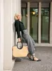 A woman leans against a building, holding a straw tote bag. Produktbild-Vorschau