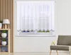 A white sheer curtain with a decorative border hangs on a window, with a wooden shelf unit and a chair visible in the room. Produktbild-Vorschau