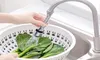 A hand is holding a flexible faucet extender over a colander filled with leafy green vegetables in a kitchen sink. Produktbild-Vorschau