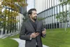 A man in a suit and scarf is standing outside a modern building, holding a smartphone and a small black case. Produktbild-Vorschau