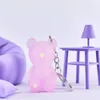 A pink bear-shaped keychain is displayed in a whimsical, pastel-colored setting with miniature furniture. Podgląd zdjęcia produktu