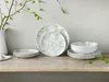 A set of porcelain plates and bowls with a botanical pattern is displayed on a wooden table, with a vase and branches in the background. Náhľad obrázku produktu