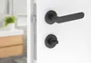 A close-up view of a black door handle and lock mechanism on a white door, with a blurred background of a bathroom vanity. Produktbild-Vorschau