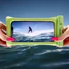 Hands holding a green waterproof phone case with a phone inside displaying a surfer on a wave, over a body of water. Produktbild-Vorschau