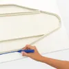A hand is marking a line on a white fabric with a blue marker, next to a beige ironing board cover. Produktbild-Vorschau