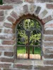 A vintage-style iron window set into a brick wall, with greenery visible through the panes. Produktbild-Vorschau