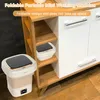 A portable mini washing machine is shown in a bathroom or laundry room setting, next to a wooden shelf unit and a white cabinet. Produktbild-Vorschau
