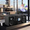 A stereo system with speakers and a CD player is displayed on a black table in front of a window with a city view. Bookshelves are visible in the background. Produktbild-Vorschau