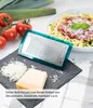 A grater is shown on a dark slate with grated cheese and a block of cheese, next to tomatoes and leeks, with a bowl of pasta in the background. Produktbild-Vorschau