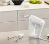 A white hand mixer with attachments sits on a kitchen counter, with kitchen cabinets and some fruit in the background. Produktbild-Vorschau