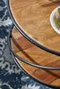 A close-up view of a round wooden coffee table with a metal frame, resting on a patterned rug. Náhľad obrázku produktu