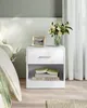 A white nightstand with a drawer and an open shelf, placed next to a bed. It holds a framed picture and a vase of flowers on top. Books are stacked in the open shelf. Náhled obrázku produktu