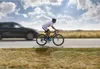 A cyclist rides on a road next to a car, with a cloudy sky and grassy fields in the background. Náhled obrázku produktu