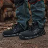 A person wearing jeans and black safety shoes standing on a dirt ground with logs in the background. Náhľad obrázku produktu