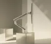 A white monitor arm is mounted on a white stand, which is placed on a metallic cube. Another metallic cube is placed to the left of the stand. The background is a light gray wall with shadows cast by sunlight. Produktbild-Vorschau